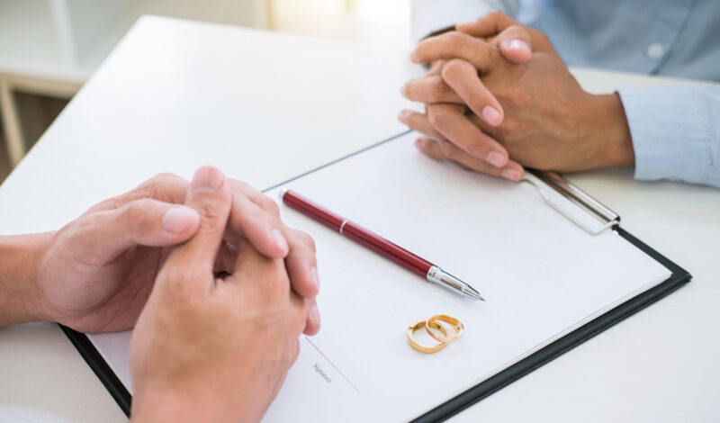 People at a desk with their hands folded over a document with wedding bands and a pen on top of it