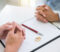 People at a desk with their hands folded over a document with wedding bands and a pen on top of it