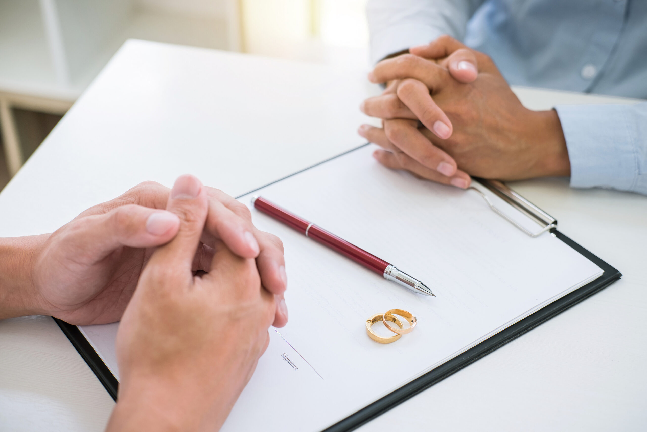 People at a desk with their hands folded over a document with wedding bands and a pen on top of it